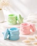 Three embossed wax melts in decorative cup-shaped containers, shown in pastel green, light blue, and soft pink, each with its matching lid leaning beside it on a bright white surface.