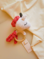 Crocheted unicorn rattle toy with red accents on a soft beige background, featuring the brand 'aera'.