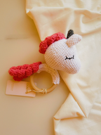 Crocheted unicorn rattle with red accents on a soft beige background