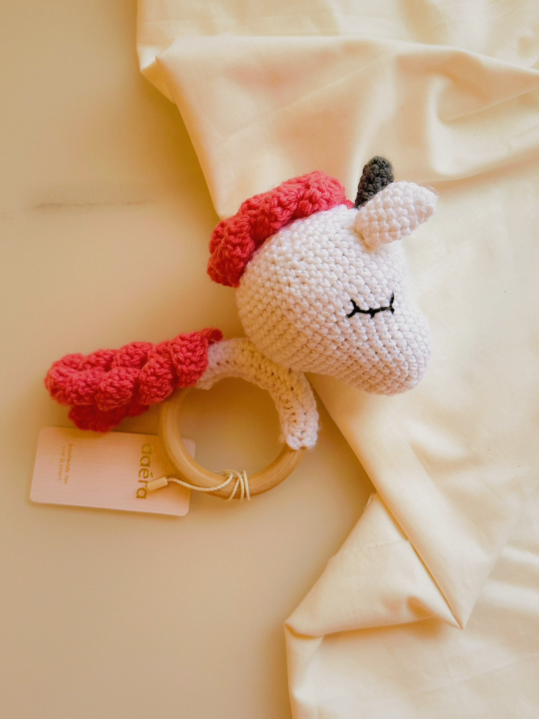Crocheted unicorn rattle with red accents on a soft beige background